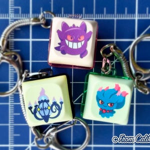 May include: Three square keychains with cartoon character designs. One keychain features a purple character with a wide grin, another has a blue and pink character, and the third has a blue and yellow design. Each keychain has a silver keyring.