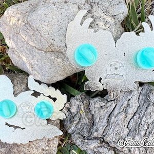 May include: Two silver-colored enamel pins with a stylized owl design, each featuring two light blue pin backs. The pins are displayed on a natural background of rocks and grass. The text "From Caliko" is visible.