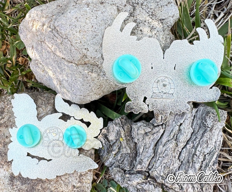 May include: Two silver-colored enamel pins with a stylized owl design, each featuring two light blue pin backs. The pins are displayed on a natural background of rocks and grass. The text "From Caliko" is visible.