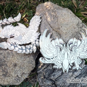 May include: Two enamel pins featuring detailed white and gray monster designs. One pin depicts a creature with a segmented body and sharp features. The other pin shows a symmetrical winged creature. The pins are displayed on a rock.