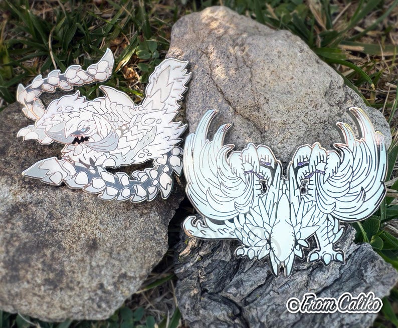 May include: Two enamel pins featuring detailed white and gray monster designs. One pin depicts a creature with a segmented body and sharp features. The other pin shows a symmetrical winged creature. The pins are displayed on a rock.