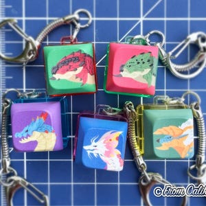 May include: Six square keychains with dragon illustrations in various colors. Each keychain has a silver-colored metal clasp and a spring coil. The dragons are depicted in red, green, blue, purple, and yellow. The background is a blue grid.