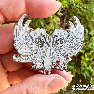 May include: A white and gold enamel pin featuring a stylized double-headed bird design with detailed wing and feather patterns. The pin has a glossy finish and is held against a blurred green background.