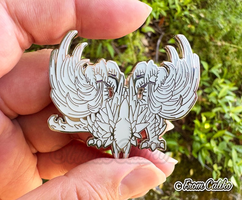May include: A white and gold enamel pin featuring a stylized double-headed bird design with detailed wing and feather patterns. The pin has a glossy finish and is held against a blurred green background.