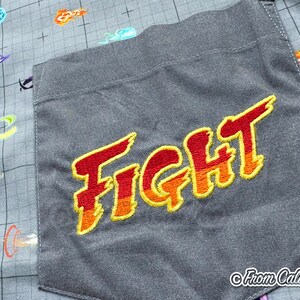 Puede incluir: Camisa gris con botones y un bolsillo bordado con la palabra "FIGHT" en rojo y naranja. La tela de la camisa tiene un patr&oacute;n de cuadr&iacute;cula y explosiones de energ&iacute;a estilizadas y coloridas.