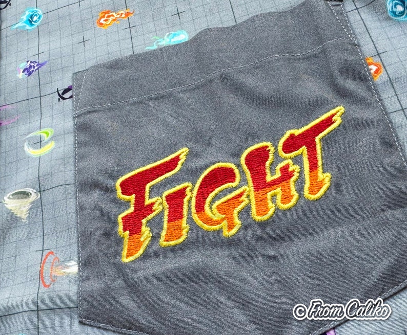 Puede incluir: Camisa gris con botones y un bolsillo bordado con la palabra "FIGHT" en rojo y naranja. La tela de la camisa tiene un patr&oacute;n de cuadr&iacute;cula y explosiones de energ&iacute;a estilizadas y coloridas.