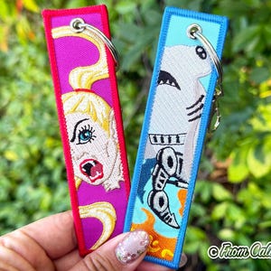 May include: Two rectangular embroidered fabric keychains with metal rings. The left keychain is pink with a blonde character face featuring blue eyes and an open mouth. The right keychain is light blue with a white shark graphic and orange flames.