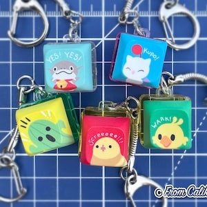 Puede incluir: Una colección de llaveros cuadrados y coloridos con varios personajes de dibujos animados y frases. Cada llavero tiene un cierre de metal plateado. Los llaveros presentan un tiburón, un gato, un cactus y pájaros. El fondo es una cuadrícula azul.