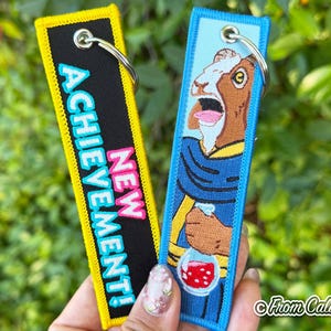 May include: Two rectangular embroidered keychains with metal rings. The left keychain is black with a yellow border and features the text NEW ACHIEVEMENT in bright blue and pink. The right keychain is light blue and features a brown goat illustration.