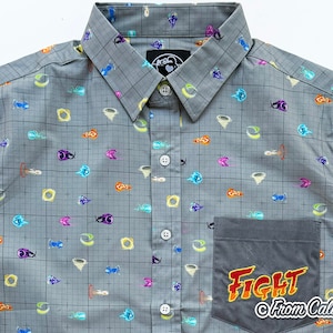 May include: Gray button-up shirt with a grid pattern and colorful cartoon graphics. The shirt features a pocket with the word "FIGHT" in orange and yellow, and the text "From Caliko".