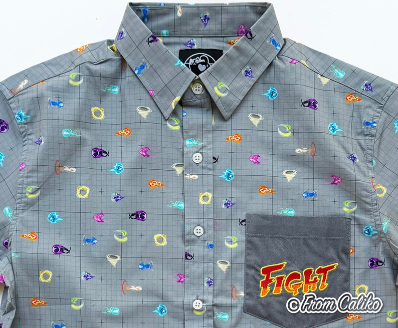 Puede incluir: Camisa gris con botones con un patr&oacute;n de cuadr&iacute;cula y gr&aacute;ficos de dibujos animados coloridos. La camisa presenta un bolsillo con la palabra "FIGHT" en naranja y amarillo, y el texto "From Caliko".