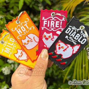 May include: Embroidered keychains in yellow, orange, red, and black, each with a cat and chili pepper design. Text on the keychains reads "MILD", "HOT", "FIRE!", and "DIABLO". Each keychain has a metal ring for attaching keys.