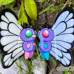 May include: Two butterfly-shaped pins with purple bodies, large iridescent wings, and cartoonish faces. One has green eyes and pink feet, the other has pink eyes and blue feet. The pins are set against a backdrop of green foliage.