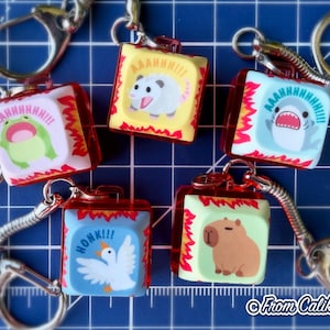 Op de afbeelding: Een verzameling sleutelhangers met cartoon dieren en tekstballonnen. Elke sleutelhanger heeft een rood frame en een zilveren sleutelring. De dieren zijn onder andere een kikker, opossum, haai, gans en capibara, elk met een unieke uitdrukking en geluidseffect.