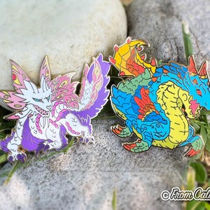 Può includere: Due spille smaltate raffiguranti creature fantasy. Una è una creatura bianca e viola con ali a farfalla. L'altra è un drago colorato con scaglie blu, gialle e rosse. Le spille sono appoggiate su una superficie di pietra.