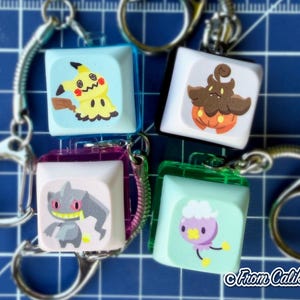 May include: Four keychains with cube-shaped charms featuring colorful cartoon characters. The charms are attached to silver keyrings. The background is a blue grid. The text "From Caliko" is in the bottom right corner.