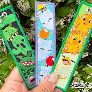 May include: Embroidered keychains with cartoon characters. One green keychain features a cactus design, a purple keychain has cat designs, and a yellow keychain has chick designs. Each keychain has a silver metal ring.