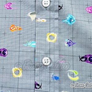 Puede incluir: Primer plano de una camisa gris con botones con un patr&oacute;n de cuadr&iacute;cula y explosiones de energ&iacute;a coloridas y de dibujos animados. Las explosiones son en tonos naranja, morado, azul y amarillo. La camisa tiene botones blancos y el texto "From Caliko".