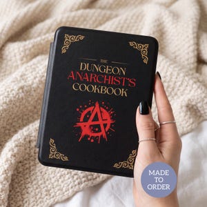 May include: A black e-reader cover featuring gold decorative corners and the text THE DUNGEON ANARCHIST'S COOKBOOK in gold and red lettering. A red anarchy symbol is printed below the title. A circular blue badge reads MADE TO ORDER.