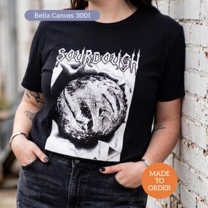 May include: Black t-shirt with the word "SOURDOUGH" in a stylized font above a black and white image of a loaf of bread. The shirt has short sleeves and a crew neck. The text "MADE TO ORDER" is in a circle on the bottom right.
