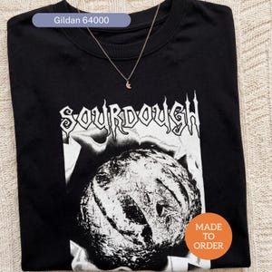 May include: Black crew neck t-shirt with a white graphic of a loaf of sourdough bread and the word "SOURDOUGH". A gold necklace with a crescent moon pendant is draped over the shirt. The shirt is labeled "Gildan 64000" and "MADE TO ORDER".