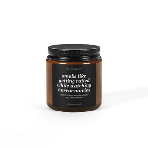 May include: A brown glass jar candle with a black lid. The label reads "smells like getting railed while watching horror movies." The text "INFUSED WITH SCREAMING FOR MULTIPLE REASONS" is also on the label. The candle is from "THE TIPSY ARTIFICER."