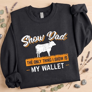 Può includere: Una felpa nera a maniche lunghe con una grafica bianca di una mucca su un prato. Il testo recita "SHOW DAD" in un carattere stilizzato, seguito da "THE ONLY THING I SHOW IS MY WALLET" in lettere bianche su uno sfondo arancione.