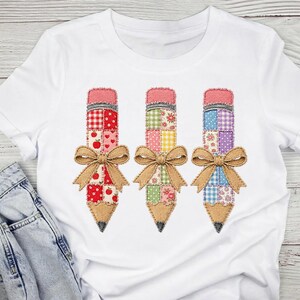 Puede incluir: Una camiseta blanca de manga corta con un gráfico de tres lápices dispuestos uno al lado del otro. Cada lápiz tiene un patrón de patchwork en tonos rojos, verdes y azules, con una goma rosa y un lazo de color beige en el centro.