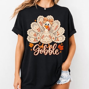 Puede incluir: Una camiseta negra de manga corta con el gráfico de un pavo con patrones de plumas de encaje, un lazo a cuadros naranja y la palabra Gobble escrita en cursiva naranja, acentuada por pequeñas hojas de otoño naranjas y estrellas.
