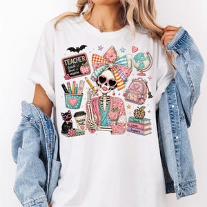 Puede incluir: Una camiseta blanca con un gráfico de un esqueleto que lleva un lazo grande estampado y gafas de sol en forma de corazón. El diseño incluye material escolar, un globo terráqueo, una mochila, un gato negro y libros con el texto Teach, Love, Inspire.