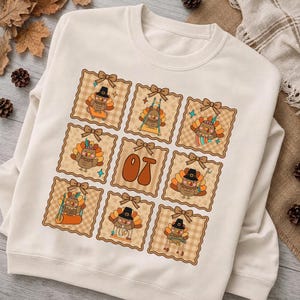 Puede incluir: Una sudadera de cuello redondo color crema con una cuadrícula de ocho marcos cuadrados con bordes festoneados y lazos. Cada marco contiene un pavo de dibujos animados en diversas actividades, mientras que el cuadrado central muestra las letras OT en naranja.