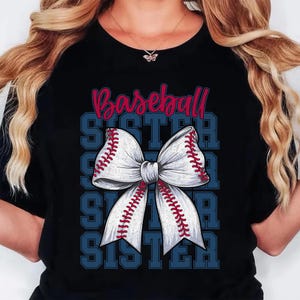 Puede incluir: Camiseta negra con un dise&ntilde;o tem&aacute;tico de b&eacute;isbol. El dise&ntilde;o incluye la palabra "Baseball" en escritura roja sobre un lazo blanco con costuras rojas, que se asemeja a una pelota de b&eacute;isbol. La palabra "SISTER" se repite en azul.