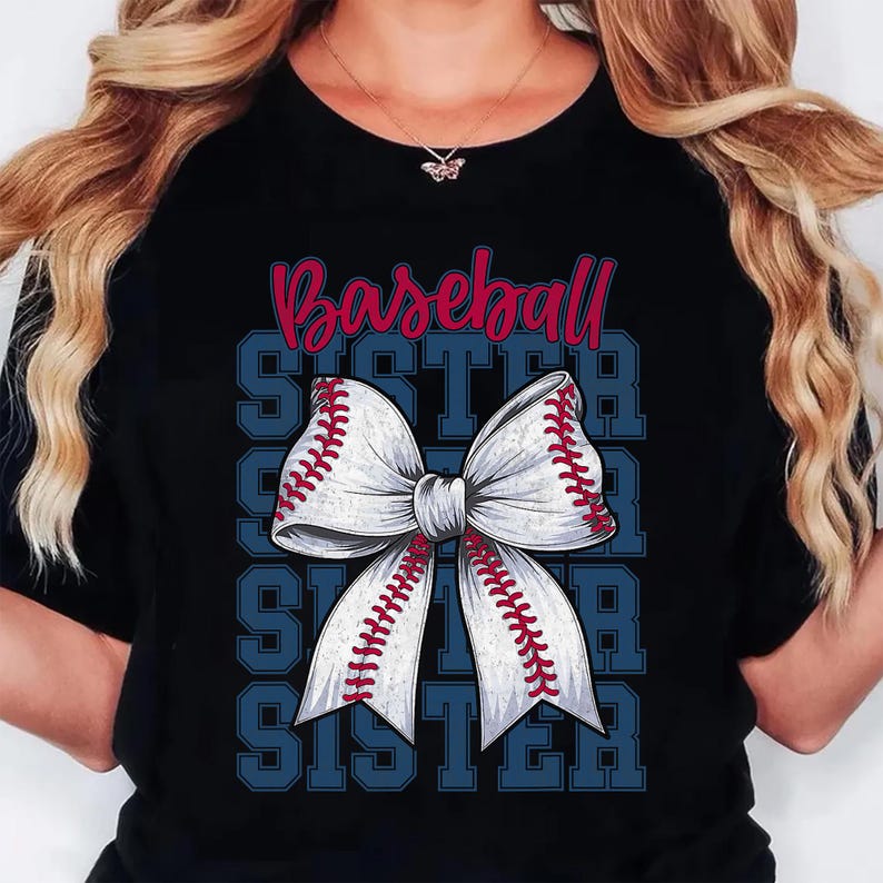Puede incluir: Camiseta negra con un dise&ntilde;o tem&aacute;tico de b&eacute;isbol. El dise&ntilde;o incluye la palabra "Baseball" en escritura roja sobre un lazo blanco con costuras rojas, que se asemeja a una pelota de b&eacute;isbol. La palabra "SISTER" se repite en azul.