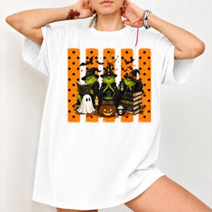 Puede incluir: Una camiseta blanca de manga corta con un gráfico de tres personajes verdes vestidos de brujas. El diseño incluye una pila de libros, un caldero, un fantasma, una calabaza con estampado de leopardo y rayas verticales naranjas con lunares negros.