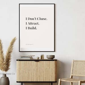 May include: A framed print with a white background and black text that reads "I Don't Chase. I Attract. I Build." The print is hanging on a white wall above a wooden cabinet with a woven rattan chair in front of it.