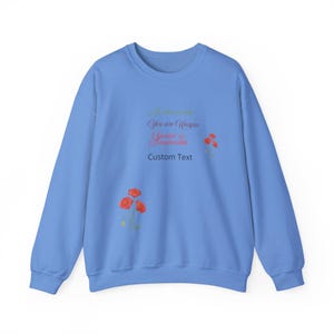 Op de afbeelding: Een lichtblauw sweatshirt met de tekst "In this world You are Unique Special & Irreplaceable" en "Custom Text" gedrukt op de voorkant. De tekst is in roze en groene lettertypen. Er zijn rode klaprozen gedrukt op de voorkant van het sweatshirt.