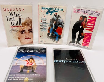 May include: Five cassette tapes with clear plastic cases. The tapes are labeled with the titles "Madonna Who's That Girl", "Beverly Hills Cop", "Pretty Woman", "Even Cowgirls Get the Blues", and "Soundtrack from Thirtysomething".