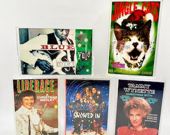 May include: A collection of five vintage cassette tapes. The tapes feature album art with various themes, including a cat in a Santa hat, a musician playing a guitar, and Christmas-themed albums. The tapes are in clear plastic cases.