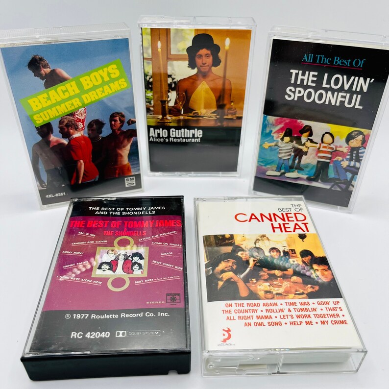 May include: A collection of five vintage cassette tapes featuring various artists, including The Beach Boys, Arlo Guthrie, The Lovin' Spoonful, Tommy James and The Shondells, and Canned Heat. The tapes are in clear plastic cases with black labels and colourful artwork.