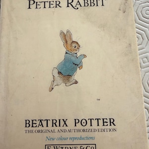 May include: A book titled The Tale of Peter Rabbit by Beatrix Potter. The cover is off-white and features a central illustration of a brown rabbit wearing a blue jacket. Text includes The Original and Authorized Edition and F. Warne & Co.