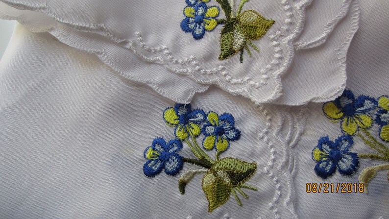 Puede incluir: Dos servilletas de tela blanca con flores bordadas azules y amarillas. Las servilletas tienen bordes festoneados y est&aacute;n superpuestas. La fecha 08/21/2018 est&aacute; escrita en la esquina inferior derecha.