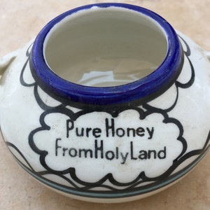May include: A small, white ceramic honey pot with a blue rim and two handles. The pot is decorated with black and blue designs and the text "Pure Honey From Holy Land".