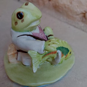 Op de afbeelding: Een keramiek beeldje van een kikker met een wit jasje en een roze vest, die een groene en oranje vis vasthoudt.