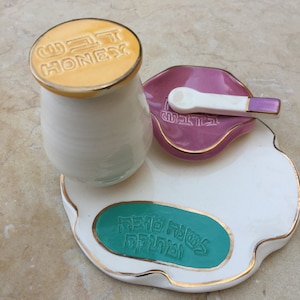 May include: A white ceramic jar with a yellow lid that says "WE'RE HONEST" on top. The jar is sitting on a white ceramic tray with a pink ceramic dish and a white ceramic spoon. The tray has a green circle with Hebrew text on it.