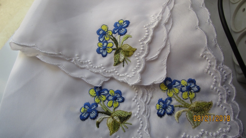 Puede incluir: Dos pa&ntilde;uelos blancos con flores bordadas azules y amarillas. Los pa&ntilde;uelos tienen un borde festoneado y est&aacute;n doblados uno encima del otro. La fecha 08/21/2018 est&aacute; bordada en la esquina inferior derecha del pa&ntilde;uelo.