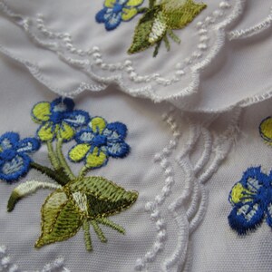 Puede incluir: Tela blanca con flores bordadas azules y amarillas. La tela tiene un borde festoneado.