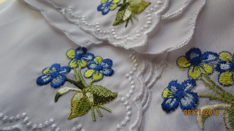 Puede incluir: Tela blanca con flores bordadas azules y amarillas. La tela tiene un borde festoneado.