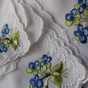 Puede incluir: Conjunto de tres pa&ntilde;uelos blancos con delicado encaje en el borde y flores bordadas en azul y amarillo. Los pa&ntilde;uelos est&aacute;n doblados y apilados uno encima del otro. La fecha 08/21/2018 est&aacute; escrita en la esquina inferior derecha de la imagen.