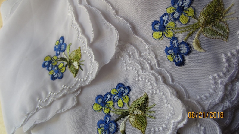Puede incluir: Conjunto de tres pa&ntilde;uelos blancos con delicado encaje en el borde y flores bordadas en azul y amarillo. Los pa&ntilde;uelos est&aacute;n doblados y apilados uno encima del otro. La fecha 08/21/2018 est&aacute; escrita en la esquina inferior derecha de la imagen.