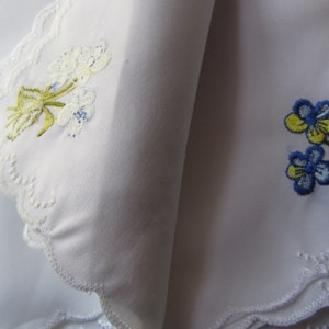 Puede incluir: Dos pa&ntilde;uelos blancos con dise&ntilde;os florales bordados. Un pa&ntilde;uelo presenta flores blancas con tallos y hojas verdes. El otro pa&ntilde;uelo presenta flores azules y amarillas con tallos y hojas verdes. Ambos pa&ntilde;uelos tienen un borde de encaje blanco.